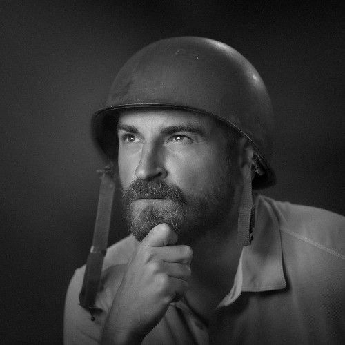 Person wearing a military helmet, with hand resting on chin, posing in a thoughtful manner.