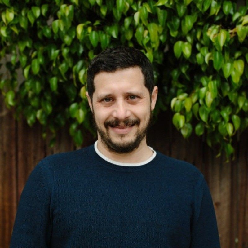 Person smiling in front of green leafy background and wooden fence.